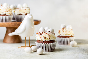 Chocolate cupcakes with cream cheese nest, egg shaped sweet candies and grated chocolate.  Bird statue. Holiday homemade dessert. Spring food background.