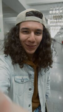 Vertical POV UGC of positive Caucasian guy with curly hair speaking about traveling around city with skateboard while recording video for vlog in underground