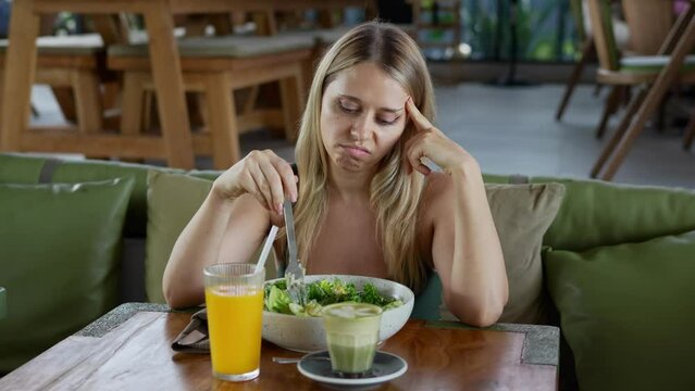 Unhappy young attractive caucasian blonde woman picking with a fork at a salad with fresh vegetables and greens sitting on green couch in the restaurant. Diet concept. Tasteless food, lack of appetite
