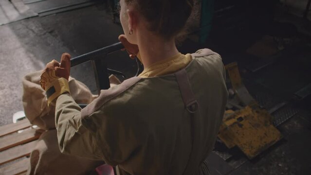 From above shot of young thin Caucasian woman in workwear carrying heavy hand pallet truck loaded with bags along dark empty factory during night shift
