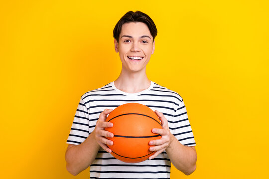 Photo of cheerful positive student with stylish hairdo dressed striped t-shirt hold bascketball ball isolated on yellow color background