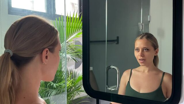 A young сaucasian blonde woman standing in front of the bathroom mirror saying affirmations aloud to raise her spirits, inspiration, and self-confidence. Psychological attitude for a positive day