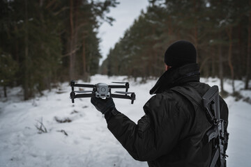 A military man with a rifle behind his back launches a drone in the forest in winter. Close up.