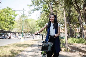 A young Asian woman commutes to work by cycling in a green city, carrying a backpack and using a...