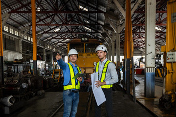 Railway technician engineer wearing safety uniform and safety helmet holding tablet and blueprint check work plan standing at site railroad station. Locomotive repairman in factory.