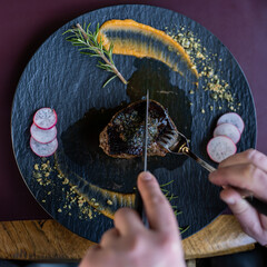 A perfectly seared steak on a slate plate, garnished with rosemary, puree swipe, and sliced radishes, with hands cutting into the juicy meat