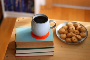 Cup of tea or coffee, pile of books, plate of cookies, reading glasses, e-reader and pen on the table. Colorful rainbow bookshelf in the background. Selective focus.