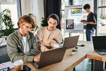 Young startup team discussing project while working on laptops in a coworking space, men in 20s