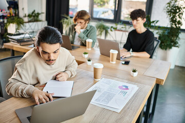 Focused young entrepreneur analyzing data on laptop and taking notes near charts on table