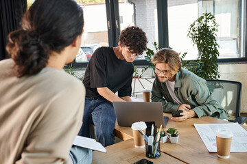Young professionals in 20s brainstorming over a laptop in a modern office near blurred coworker