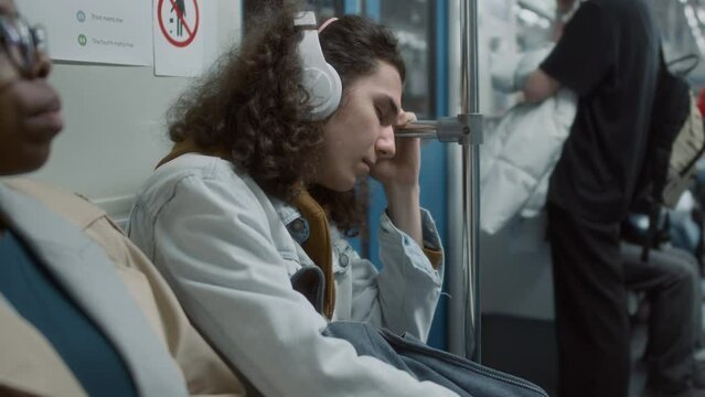 Side chest up footage of tired Caucasian young man in headset sleeping with backpack in transit subway train holding onto handrail while riding home after hard workday