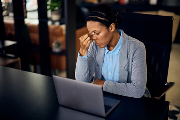 A black businesswoman having a headache, feeling tired while working online in her office.