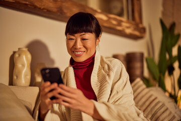 A smiling Asian woman sitting at home and using a mobile phone.