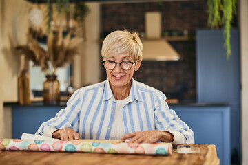 A happy senior woman using a wrapping paper and packing a gift for someone, at home.