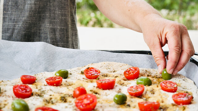 Woman preparing traditional Italian focaccia puts olives and tomatoes on top of the dough. Cooking outside in the garden with fresh ingredients.