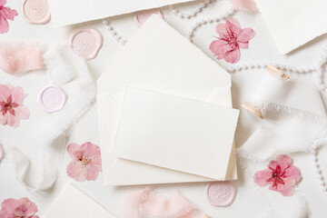 Fototapeta na wymiar Card and envelope near pink decorations, seals and silk ribbons on white table top view, wedding mockup
