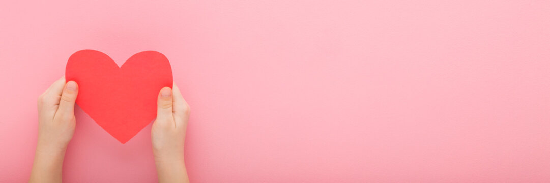 Little child hands holding red paper heart shape on pink table background. Pastel color. Love, health and hope concept. Closeup. Wide banner. Empty place for text. Point of view shot. Top down view.