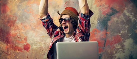 A young man in his 20s is sitting at a laptop with his arms in the air, displaying a gesture of celebration and success. He appears enthusiastic and engaged in his work. - obrazy, fototapety, plakaty