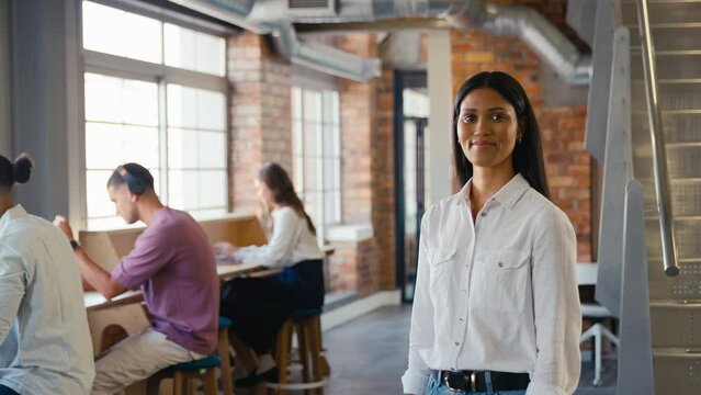 Portrait of smiling young businesswoman working in modern open plan office turning to look at camera and folding arms with colleagues in background - shot in slow motion