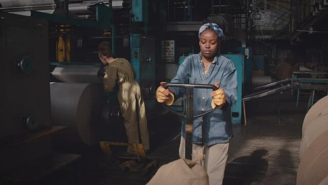 Lowkey shot of young African American female industrial factory worker pulling hand pallet truck loaded with heavy bags during work shift with colleague repairing machine equipment