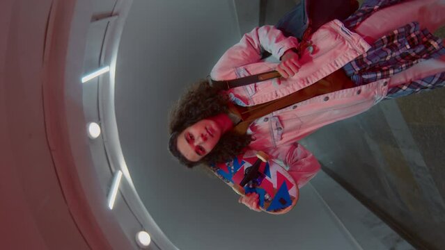 Rotating push in portrait of Caucasian curly-haired skater posing with bright skateboard in empty long tunnel or underpass with red neon light and looking at camera
