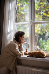 Lonely upset young woman leaning on windowsill near cat need psychologies help. Outcast by friends...