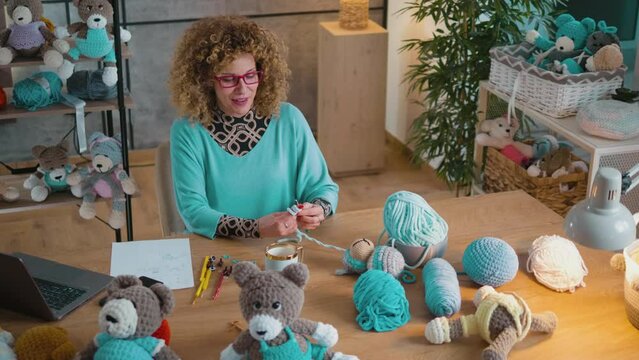 Smiling woman chatting in video call on laptop while crocheting stuffed toys at home, having her own small business