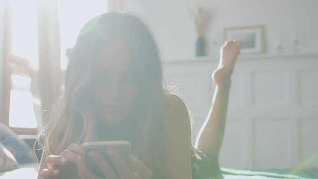 young cheerful smiling woman with long hair lies on bad at sun light, relaxing at daytime, used apps in mobile phone, online shopping, scrolling and swiping.