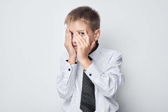 Scared young boy covering face with hands, isolated on a gray background.