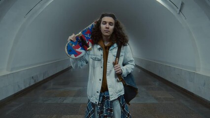 Dolly zoom portrait of Caucasian cool gen Z guy in skater style clothes posing with vividly colored board in empty long underpass and looking at camera