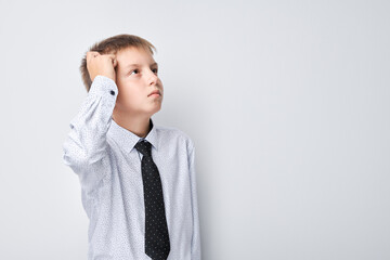 Pensive young boy in shirt and tie scratching head, looking away on gray background.