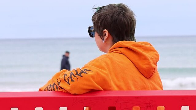 Mujer sentada en banco rojo de la Playa de San Juan mirando a c&aacute;mara y volteandose mientras un paseante no reconocible pasea por la orilla del mar, Alicante, Espa&ntilde;a