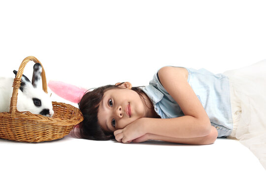 Cute little girl wearing pink bunny ears lying down on floor with rabbit in basket. Joyful kid and bunny celebrating Easter day on white background. Happy child on Easter holiday celebration.