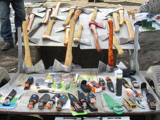 A man sells axes and knives at a flea market. The goods are laid out directly on the ground