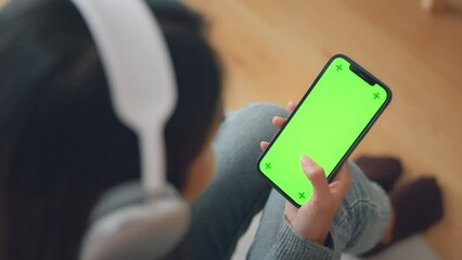 Woman with headphones holding mockup blank green screen mobile smartphone while listening online music media entertainment relaxation on sofa at home, Green empty space for advertise text
