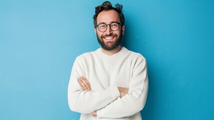 An attractive young man holding a camera smiling with crossed arms and a beard wearing a casual sweater and glasses against a blue background. This is a positive person with a happy face. - obrazy, fototapety, plakaty