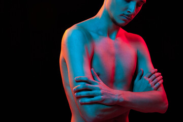 Cropped portrait of bare-chested young handsome man posing crossing hands in neon light against...