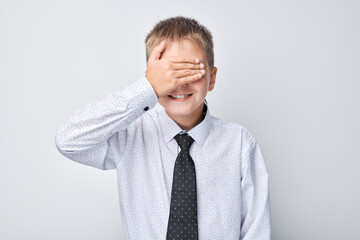Smiling boy in shirt and tie covering his eyes with hand, playing hide and seek on plain background