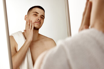 Young man in bathroom looking to mirror and touching his face, examining his stubble after shaving....