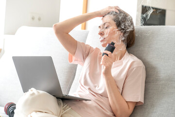 Young woman using disposable electronic cigarette during work on laptop at home