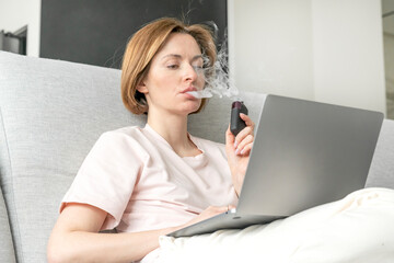 Young woman using disposable electronic cigarette during work on laptop at home