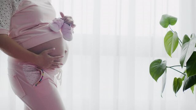 Pregnant woman stroking belly near window indoors. Adult expectant mother standing in light room. Future mom near window at home strokes touches her big belly. White pregnant woman. Slow motion