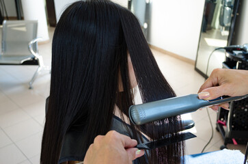 Planchado o alisado de cabello negro a clienta en Salón de Belleza con plancha eléctrica de pelo y peine. Servicio de peluquería profesional. 