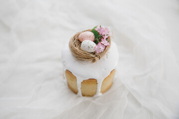 Easter cake with white glaze with a decorative bird's nest with eggs and flowers on a wooden board and white gauze. Copy space. 