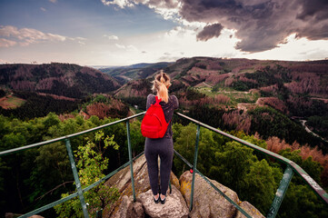 A hiker stands at a mountain lookout point, hands raised in a gesture of freedom, with a dramatic sunset sky above the vast forest landscape