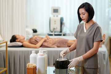Waxing salon worker preparing supplies for procedure, client waiting in background
