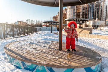 A little todd girl in a red down jacket with fur on a playground in winter on a sunny day. Saint...
