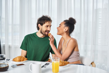 jolly african american woman in homewear feeding croissant to her handsome loving boyfriend