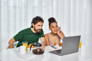 Fototapeta na wymiar attractive jolly multicultural couple in cozy homewear watching movies on laptop during breakfast