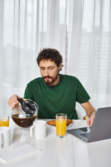 good looking man in comfy homewear pouring some coffee during breakfast with laptop on table